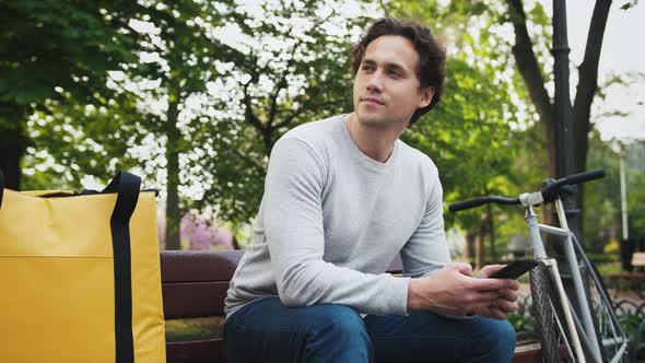 Portrait of Caucasian Delivery Man Sitting on Bench in Central Park and Using Smartphone with Yellow alt