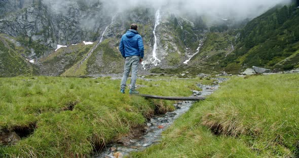 Hiker or Backpacker Trekking in the Mountains Standing in Front of Alpine Valley with Waterfall alt