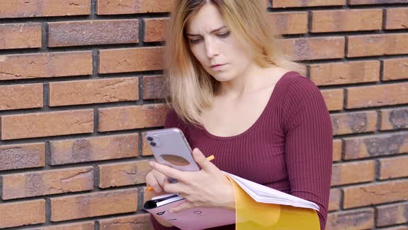 Young Female with Notebook Looks at Phone and Writes Near Wall