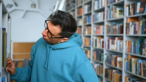 A Young Millennial Student with Glasses Selects an Interesting Book on the Bookshelves in a Public