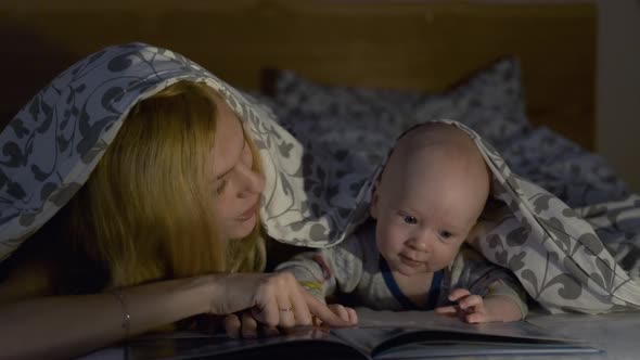 mom and baby read a book while lying under the covers