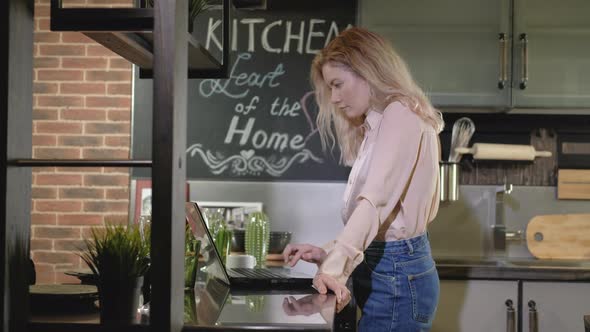 Nice young girl in kitchen and uses laptop to check mail or order goods online alt