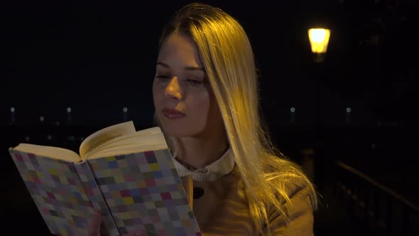 A Young Beautiful Woman Reads a Book in an Urban Area at Night - Streetlight in the Background alt