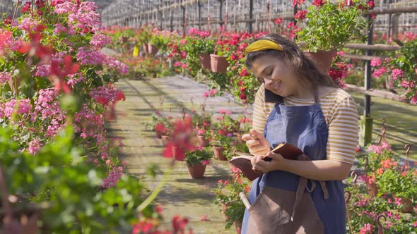 Woman Talks on Smartphone Writing in Notebook in Greenhouse