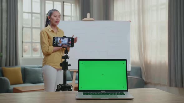 Asian Female Teacher With Green Screen Laptop Shoots Video By Smartphone While Teaching English alt