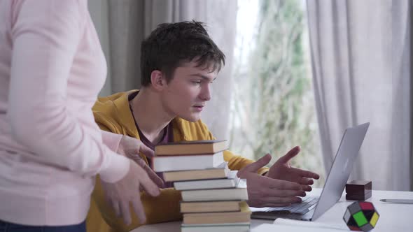 Side View of Young Caucasian Boy Gaming As Unrecognizable Woman Putting Stack of Books on Table and alt