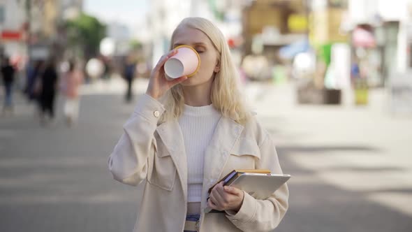 A Young Blonde Woman Stands in the Middle of the Street with Books and a Tablet and Drinks Delicious