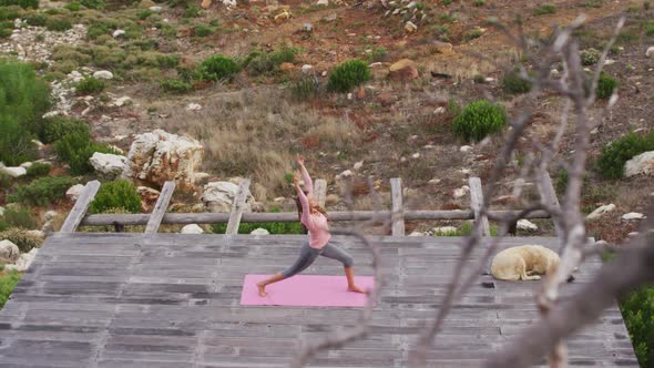 Caucasian woman practicing yoga outdoors, stretching standing on deck in rural mountainside setting alt