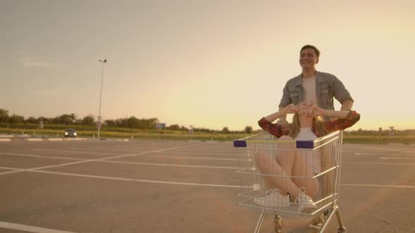 Side View of a Young Female and Male Having Fun Outdoors on Shopping Trolleys alt
