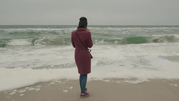 Girl in a Marsal Coat Looks Into a Smartphone Against the Background of a Cold Winter Sea with