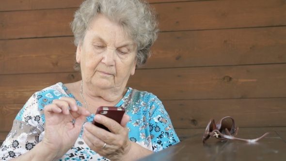 Old Woman Sitting At The Table Holds a Smartphone