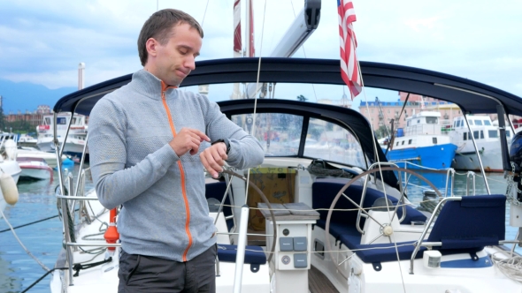 Handsome Young Man Standing Near His Yacht. He Checks Messages On The Smart Watch And Meets Friends