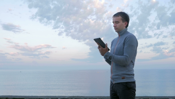 A Man Stands Near The Ocean With The Tablet. He Checks Messages On The Social Network