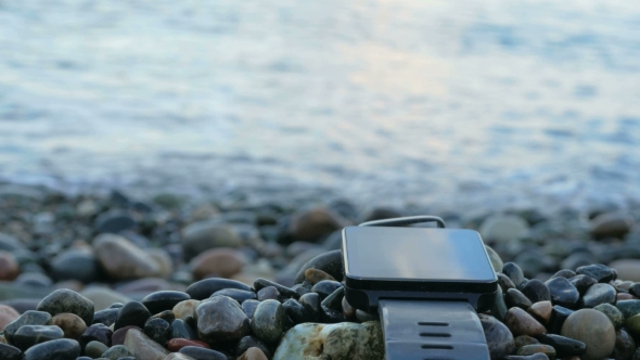 Blurred Smart Watches Are Near The Pebble Beach Of The Ocean. The Clock Reflects The Sky Clouds. .