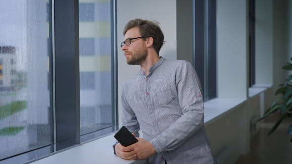 Young Man Standing In The Corridor, Holding a Smart Phone And Looking Out The Window. alt