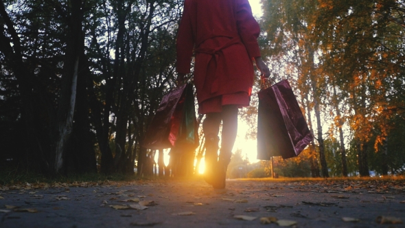 Young Woman In A Raincoat With Shopping Bags Goes In The Park Trough The Sun During Sunset
