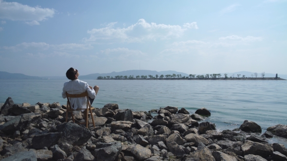 Man In a White Jacket Sitting On a Chair On The Riverbank And Enjoying The View alt