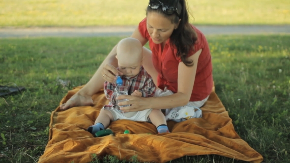 Mom Playing With Baby Son In The Park. They Sit On a Blanket On The Grass And Smiling