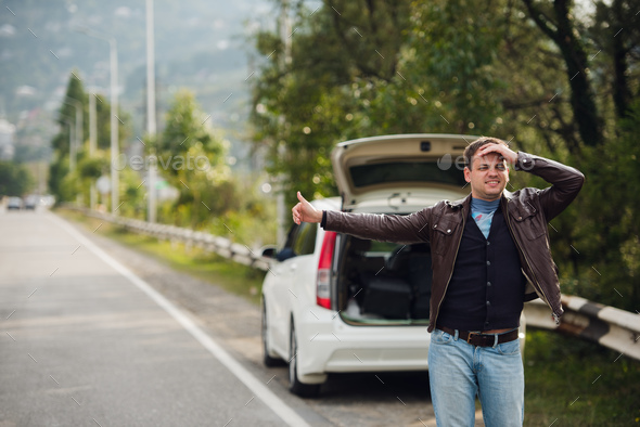 Hitchhiking - Need a drive. Young man on the road with his hand raised in front of car