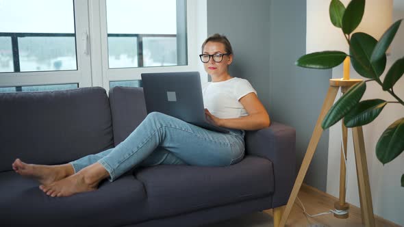 Woman with Glasses is Sitting on the Couch and Working on a Laptop alt