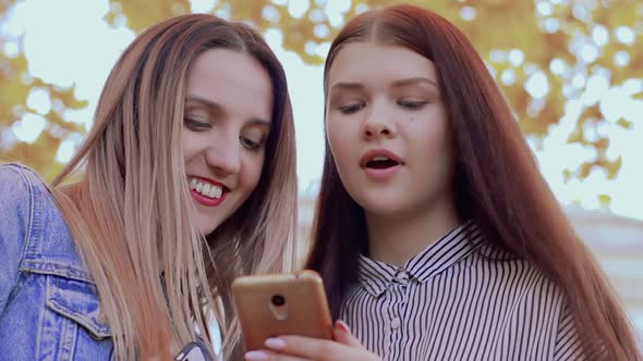 Two Smiling Girls Are Looking at Phone in the Autumn Park alt