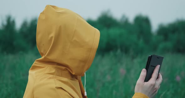 Man in a Raincoat with a Hood is in Nature and Looks at a Map in His Phone