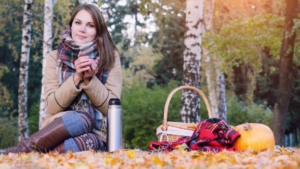Young Beautiful Woman Sitting On Picnic Drinking Hot Tea From a Thermos In Autumn Park. Girl Sitting