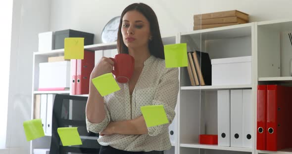 Businesswoman with Cup in Hands Considering Stickers on Glass in Office