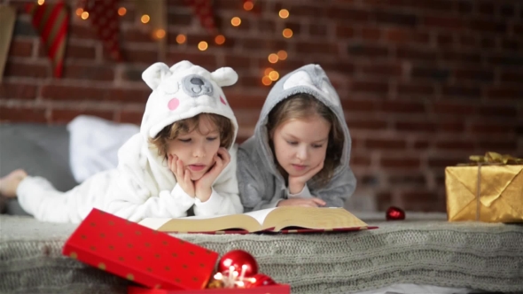 Two Little Cute Siblings Reading a Book In Bed Near Christmas Tree With Lights And Illumination alt