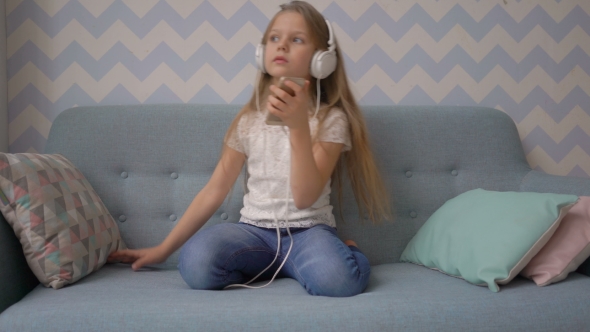 Little Girl In Headphones Is Listening To Music From Smatphone While Sitting On Sofa At Home