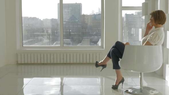 Stylish Woman Sits On The White Chair And Haves Phone Call Against Window With Cityscape alt
