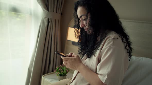 Cheerful Young Caucasian Woman Using Smartphone While Sitting on Bedroom alt
