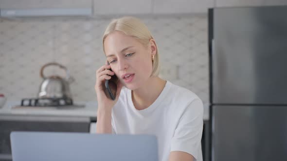Attractive Blonde Woman Talks on Phone While Working on Laptop in Kitchen