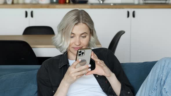 Young Caucasian attractive blonde woman looks at the screen, texting a message on the smartphone alt