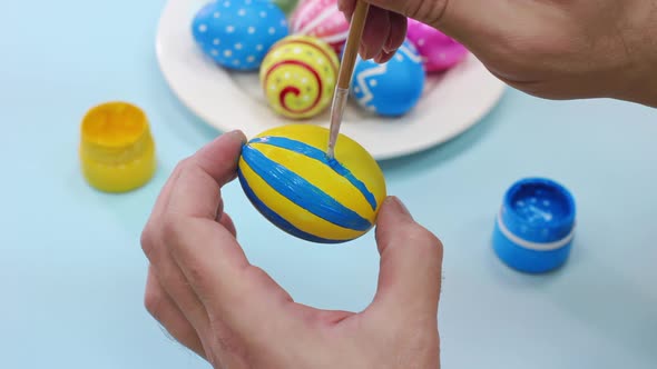 Happy Easter Close Up. A white man paints blue lines on a yellow Easter egg with a brush