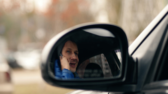 Young Man Sitting in a Car and Talking on the Phone, Shot Through the Mirror Car