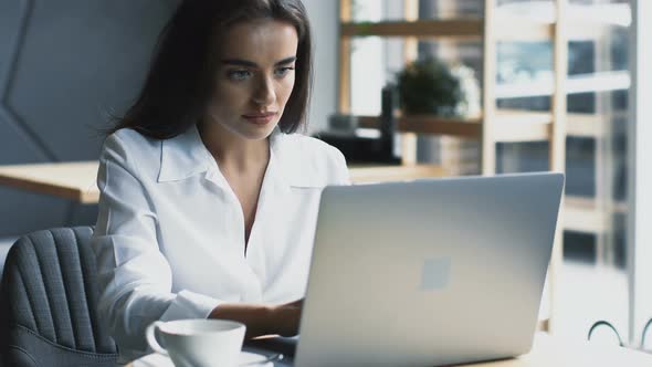 Confident and Beautiful Freelance Woman Working on Laptop in Cafe with Cup of Coffee alt