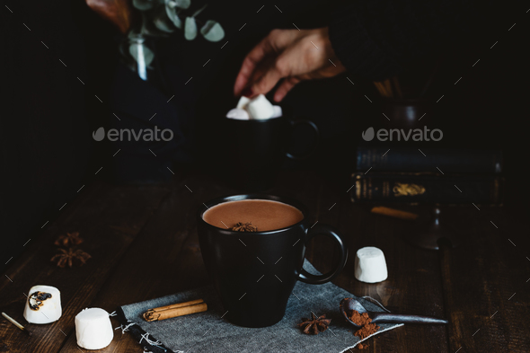 Comforting Mug of Hot Chocolate on Rustic Table with Female Hand Picking Marshmallows in Background