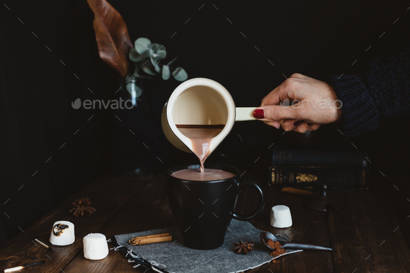 Female Pouring Hot Chocolate Drink from Milk Pan into Black Mug on Rustic Table - Stock Photo - Images