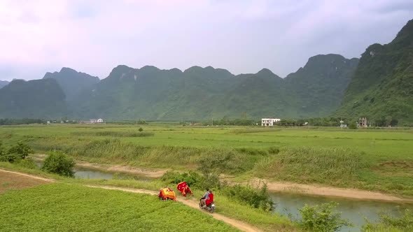 Man and Woman Ride Scooter on Narrow Ground Road Along River alt