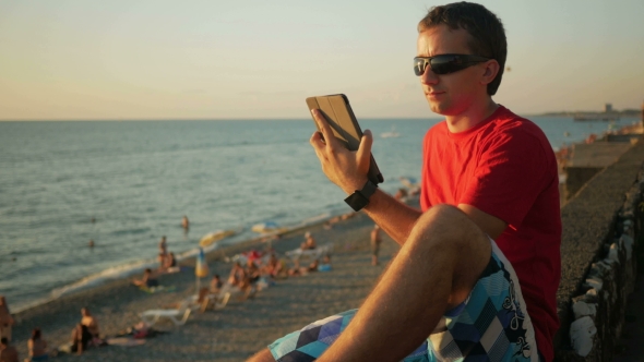 Young Handsome Man in Sunglasses Relaxing Near the Sea Beach with Tablet Computer. He Checks