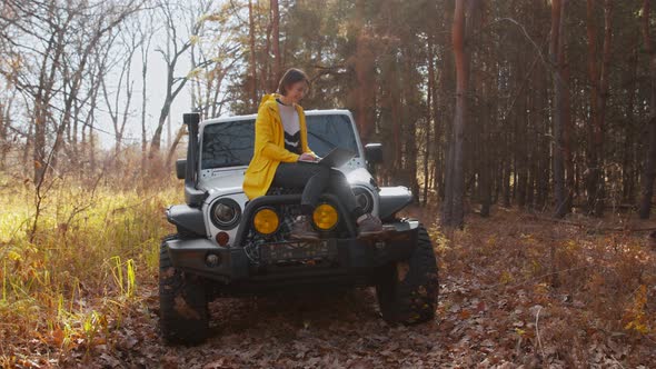 Young Happy Woman in Yellow Jacket Sits on Car and Works with Notebook in Autumn Forest