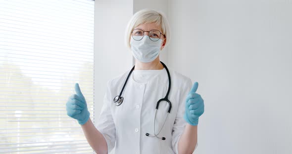 Mature woman doctor receive good results show thumb-up, wear face mask and gloves. alt