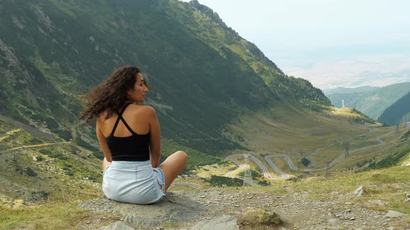A cinematic view from behind of a young woman sitting on a cliff face looking out over the mountains