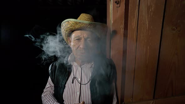 An elderly farmer in a straw hat smokes a cigar outside the ranch. The man smokes a cigar alt