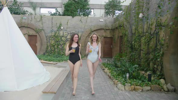 Two young attractive woman walking in one-piece swimsuit drinking healthy detox orange juice in a lu alt