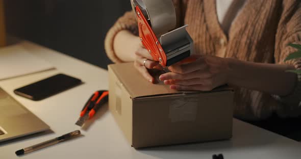 Woman Is Sealing Cardboard Box By Rollon Tape Dispenser on Her Working Table at Evening Delivery alt