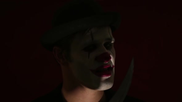 Terrible Man in the Makeup of a Clown Looks at the Camera, Laughs and Licks a Knife Blade. Terrible
