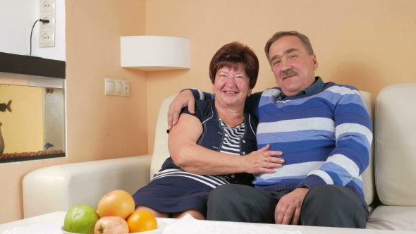 Happy Senior Man Embracing His Beloved Wife on a Sofa in a Beautiful House. They Talk and Laugh