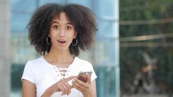 Young Millennial African American Woman Looking in Smartphone, Looking Surprised at Camera Raise alt